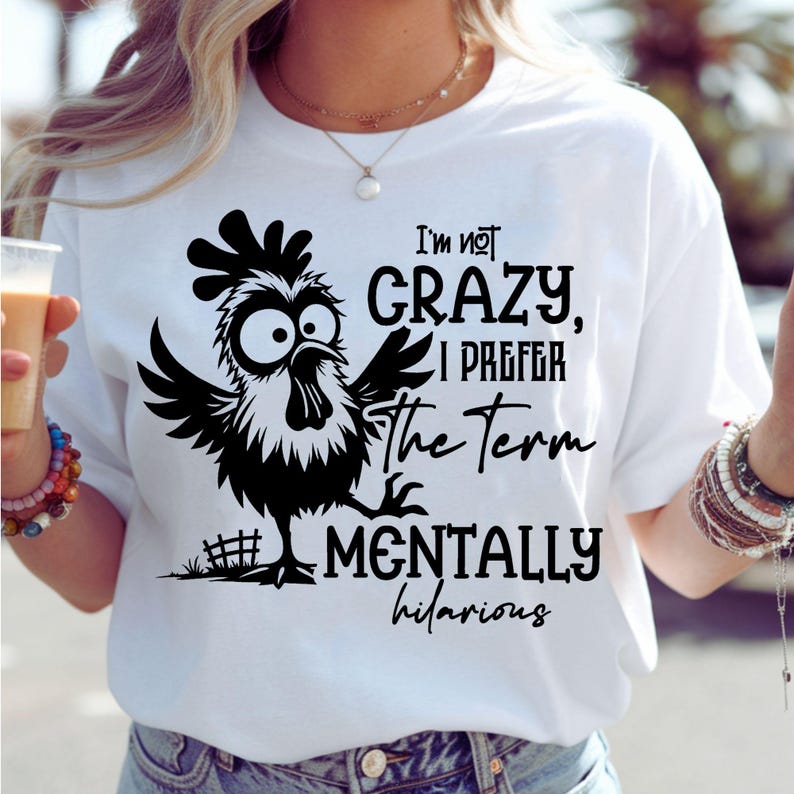 Może przedstawiać: Biała koszulka z czarnym rysunkowym kogutem i napisem "I'm not crazy, I prefer the term mentally hilarious." Kogut ma duże oczy i rozłożone skrzydła. Koszulka jest w stylu casual.