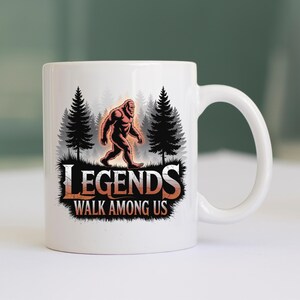 Puede incluir: Taza de cer&aacute;mica blanca con una ilustraci&oacute;n de Bigfoot frente a un bosque. La taza muestra el texto "LEGENDS WALK AMONG US" en una fuente audaz. El dise&ntilde;o incluye tonos naranja, negro y gris.