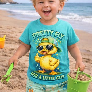 May include: A young child on a beach wearing a turquoise t-shirt with the text "PRETTY FLY FOR A LITTLE GUY" and a graphic of a duck wearing sunglasses and a hat. The child is holding a green bucket and a green shovel.