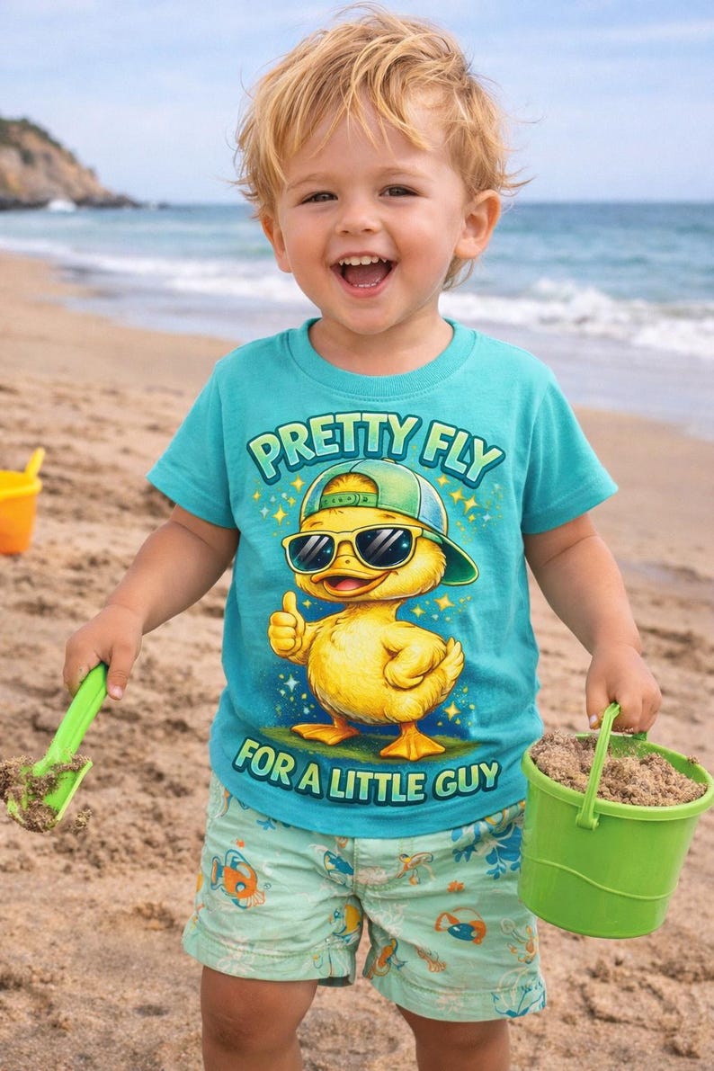 May include: A young child on a beach wearing a turquoise t-shirt with the text "PRETTY FLY FOR A LITTLE GUY" and a graphic of a duck wearing sunglasses and a hat. The child is holding a green bucket and a green shovel.