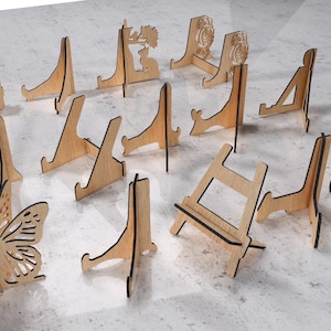 May include: A collection of wooden display stands in various designs. Some stands feature decorative cutouts, including a butterfly, a floral pattern, and a deer. The stands are made of light-colored wood with dark edges.