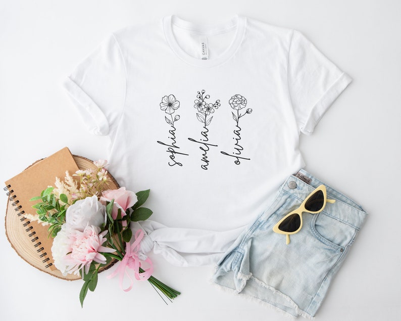 May include: White t-shirt with a floral design and the names "Sophia", "Amelia", and "Olivia" written vertically.