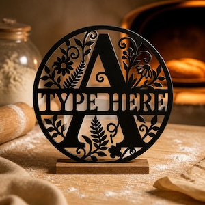 May include: A circular black metal sign featuring a large capital letter A as the central element, with the text TYPE HERE cut out across the middle. The design is surrounded by intricate floral and leaf patterns and stands on a wooden base.