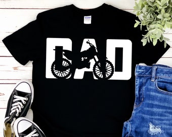 May include: Black t-shirt with the word "DAD" in large white letters. The letter "A" is replaced by a silhouette of a motorcycle. The shirt is paired with blue jeans and black sneakers.