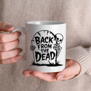 K&ouml;nnte beinhalten: Wei&szlig;e Keramiktasse mit einer schwarzen Grafik eines Skeletts, das aus einem Grabstein auftaucht. Der Text "BACK FROM THE DEAD" ist auf dem Grabstein abgebildet. Die Tasse hat einen wei&szlig;en Henkel und wird von einer Person gehalten.