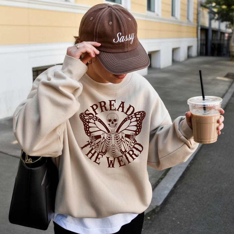 K&ouml;nnte beinhalten: Beiges Sweatshirt mit einem Skelett-Schmetterling-Motiv und dem Text "SPREAD THE WEIRD". Eine braune Baseballkappe mit dem Wort "Sassy" wird getragen. Eine schwarze Tragetasche und ein Kaffeegetr&auml;nk sind ebenfalls zu sehen.