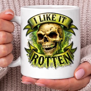 May include: White ceramic mug featuring a detailed illustration of a skull with the text "I LIKE IT ROTTEN" in a green banner. The skull has a decaying appearance with green accents, creating a spooky aesthetic.