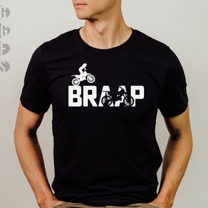 Op de afbeelding: Een zwart T-shirt met een witte afbeelding van een motorcrosser die een stunt uitvoert met het woord "BRAP" eronder.