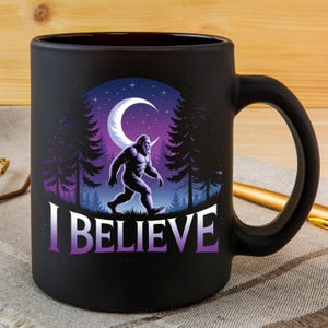 Puede incluir: Taza de cer&aacute;mica negra con una ilustraci&oacute;n de Bigfoot caminando en un bosque bajo una luna creciente y un cielo estrellado. Las palabras "I BELIEVE" est&aacute;n impresas en degradado p&uacute;rpura.