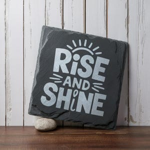 May include: A black slate sign with the words "Rise and Shine" in silver lettering. The sign is decorated with a sunburst design.