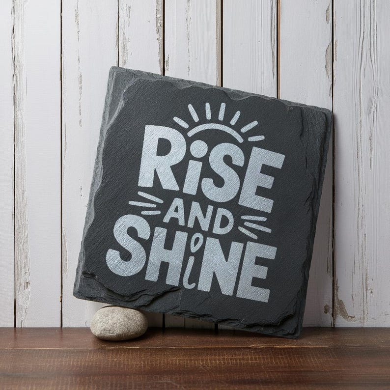 May include: A black slate sign with the words "Rise and Shine" in silver lettering. The sign is decorated with a sunburst design.