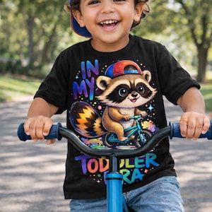 May include: A smiling child wearing a baseball cap and black t-shirt with a raccoon graphic riding a blue bicycle. The shirt reads "IN MY TODDLER ERA". The child is riding on a paved path.