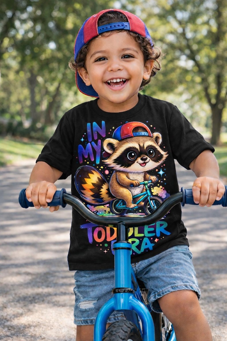 May include: A smiling child wearing a baseball cap and black t-shirt with a raccoon graphic riding a blue bicycle. The shirt reads "IN MY TODDLER ERA". The child is riding on a paved path.