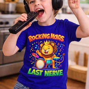 May include: A young child wearing headphones and a blue t-shirt with the text "Rocking Mods" and "Last Nerve". The shirt features a cartoon lion playing drums. The child is holding a remote control and making a rock and roll hand gesture.