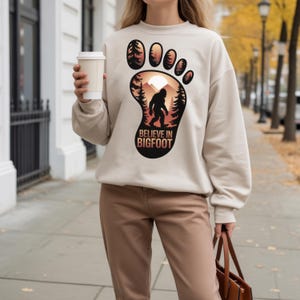 Puede incluir: Sudadera beige con un dise&ntilde;o de huella de Bigfoot. El dise&ntilde;o incluye una silueta de Bigfoot, &aacute;rboles y una escena de monta&ntilde;a, con el texto "BELIEVE IN BIGFOOT". La sudadera es de color neutro y tiene mangas largas.