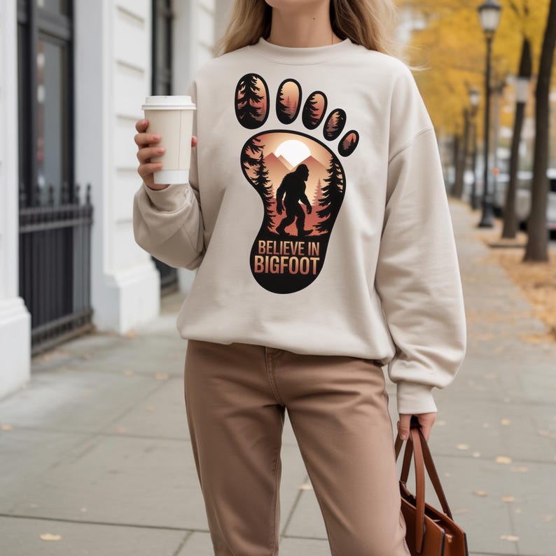 Puede incluir: Sudadera beige con un dise&ntilde;o de huella de Bigfoot. El dise&ntilde;o incluye una silueta de Bigfoot, &aacute;rboles y una escena de monta&ntilde;a, con el texto "BELIEVE IN BIGFOOT". La sudadera es de color neutro y tiene mangas largas.