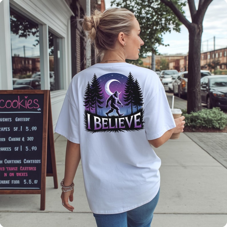 Puede incluir: Camiseta blanca con un dise&ntilde;o de Bigfoot. El dise&ntilde;o presenta una silueta de Bigfoot caminando frente a una luna y &aacute;rboles, con el texto "I BELIEVE" debajo. La camiseta es de estilo casual.