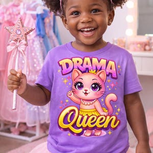May include: A young child wearing a purple t-shirt with a "Drama Queen" graphic featuring a cat, a pink tutu, and a pink crown. She is holding a pink star-shaped wand. The background includes pink dresses.