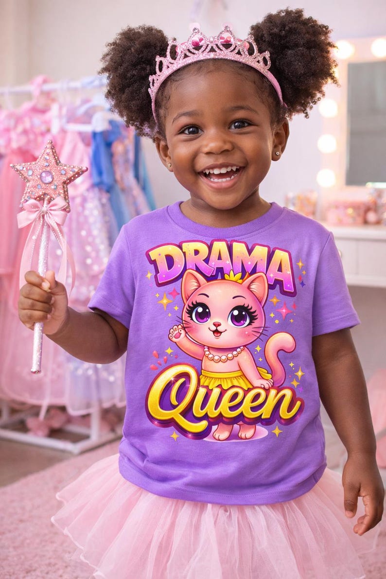 May include: A young child wearing a purple t-shirt with a "Drama Queen" graphic featuring a cat, a pink tutu, and a pink crown. She is holding a pink star-shaped wand. The background includes pink dresses.