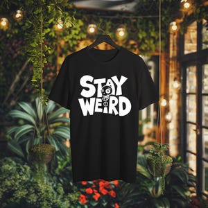 K&ouml;nnte beinhalten: Schwarzes T-Shirt mit dem wei&szlig;en Schriftzug "STAY WEIRD" und einem Totenkopf-Motiv. Das T-Shirt h&auml;ngt an einem schwarzen Kleiderb&uuml;gel vor einem Hintergrund aus Gr&uuml;npflanzen und Lichterketten.