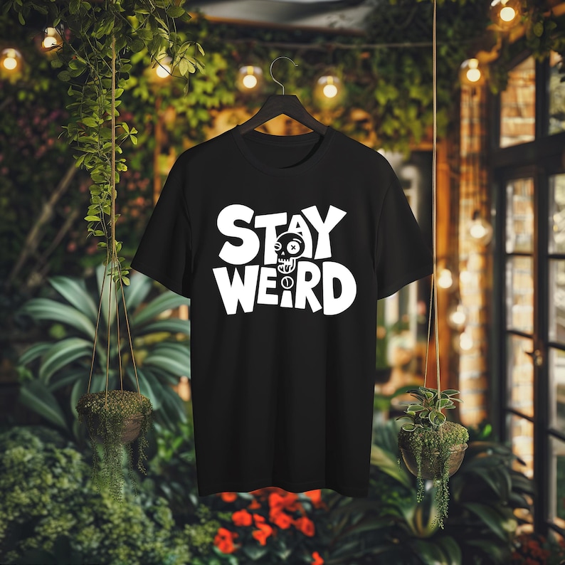 K&ouml;nnte beinhalten: Schwarzes T-Shirt mit dem wei&szlig;en Schriftzug "STAY WEIRD" und einem Totenkopf-Motiv. Das T-Shirt h&auml;ngt an einem schwarzen Kleiderb&uuml;gel vor einem Hintergrund aus Gr&uuml;npflanzen und Lichterketten.