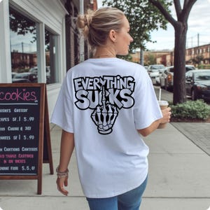 K&ouml;nnte beinhalten: Wei&szlig;es T-Shirt mit dem Text "EVERYTHING SUCKS" in fetter, umrandeter Schrift und einer Grafik einer Skeletthand, die den Mittelfinger zeigt. Das Shirt hat eine lockere Passform.