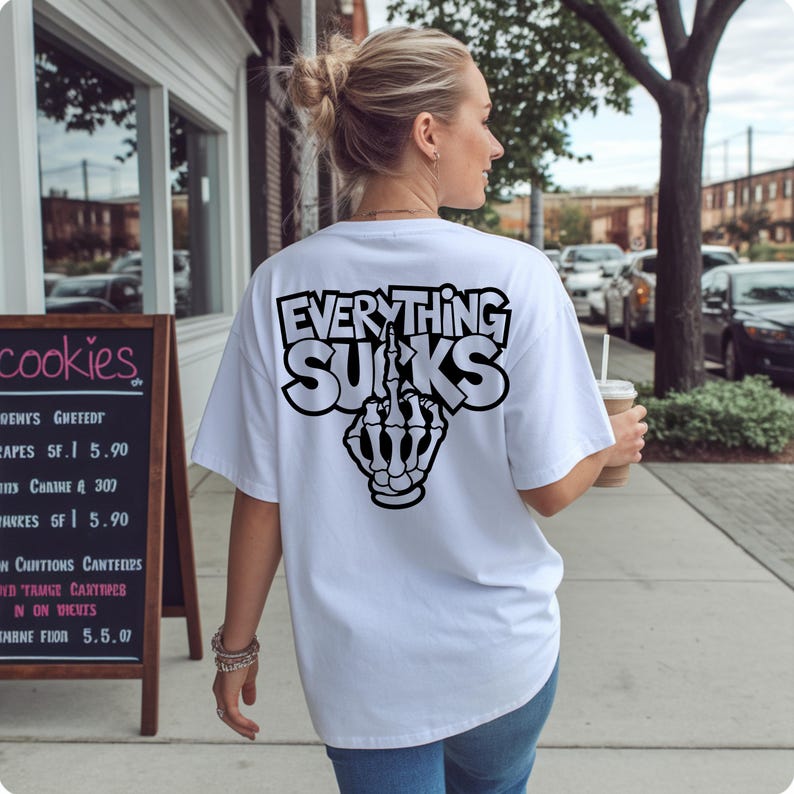 K&ouml;nnte beinhalten: Wei&szlig;es T-Shirt mit dem Text "EVERYTHING SUCKS" in fetter, umrandeter Schrift und einer Grafik einer Skeletthand, die den Mittelfinger zeigt. Das Shirt hat eine lockere Passform.