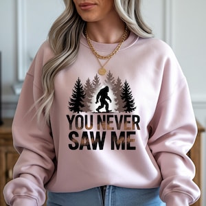 Puede incluir: Sudadera rosa claro con un dise&ntilde;o de Bigfoot. El dise&ntilde;o presenta una silueta negra de Bigfoot frente a un bosque. El texto "YOU NEVER SAW ME" est&aacute; impreso en negro y marr&oacute;n. La sudadera es de cuello redondo.