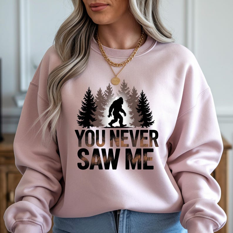 Puede incluir: Sudadera rosa claro con un dise&ntilde;o de Bigfoot. El dise&ntilde;o presenta una silueta negra de Bigfoot frente a un bosque. El texto "YOU NEVER SAW ME" est&aacute; impreso en negro y marr&oacute;n. La sudadera es de cuello redondo.
