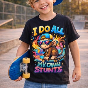 May include: A young child wearing a black t-shirt with the text "I DO ALL MY OWN STUNTS" and a graphic of a monkey on a skateboard. The child is also wearing a blue and yellow baseball cap and holding a blue skateboard.