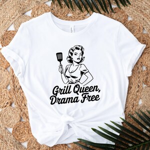 May include: White t-shirt with a black and white illustration of a woman holding a spatula. The text "Grill Queen, Drama Free" is printed in black below the illustration.