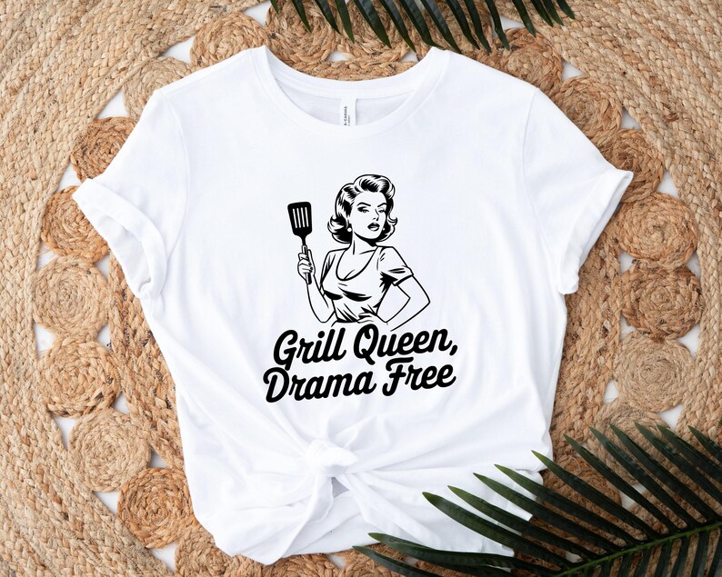 May include: White t-shirt with a black and white illustration of a woman holding a spatula. The text "Grill Queen, Drama Free" is printed in black below the illustration.
