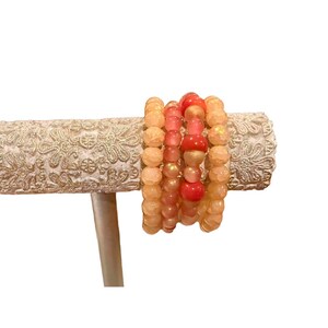 May include: Several beaded bracelets in peach, coral, and gold tones. The bracelets are presented on a decorative, off-white, patterned stand. The beads are of different sizes and shapes, creating a layered effect.