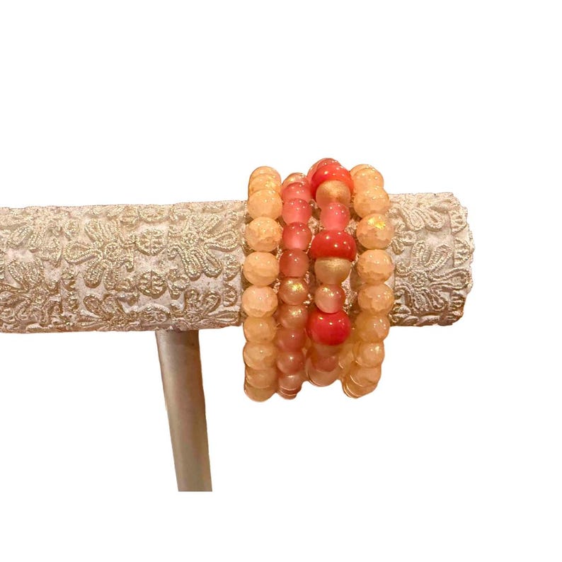 May include: A collection of beaded bracelets in shades of peach, coral, and gold. The bracelets are displayed on a decorative, off-white, patterned stand. The beads vary in size and shape, creating a layered effect.