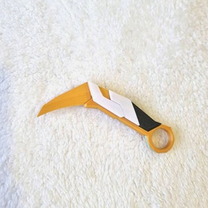 May include: A curved, yellow and white plastic prop knife with a black handle. The knife has a rounded end and a sharp point.