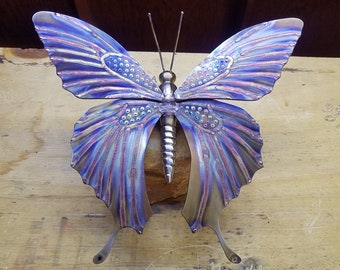 May include: A metal butterfly sculpture with iridescent blue and purple wings. The wings have a textured pattern and are decorated with small, round, silver accents.