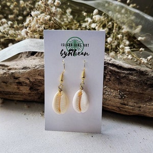 May include: A pair of gold shell earrings with a white shell pendant. The earrings are on a white card with the text "Island Girl Art by Rhean".