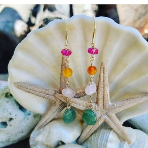 May include: Gold earrings with a layered design featuring pink, orange, and green gemstones. The earrings are displayed on a seashell and starfish background.