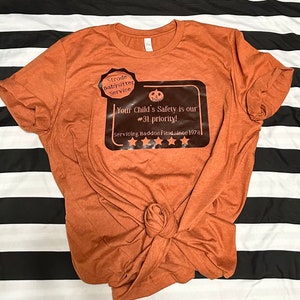 May include: A brown t-shirt with a black and orange graphic that says "Strode Babysitter Service" and "Your Child's Safety is our #31 priority! Servicing Haddonfield since 1978".