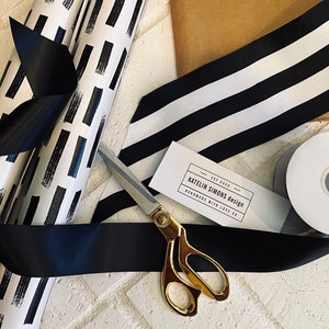 Op de afbeelding: Zwart-wit gestreept lint, zwart lint, gouden schaar en een rol zwart-wit gepatroneerd inpakpapier. Een klein wit kaartje met de tekst "EST 2020 KATELIN SIMONS design HANDMADE WITH LOVE XO" ligt op het lint.
