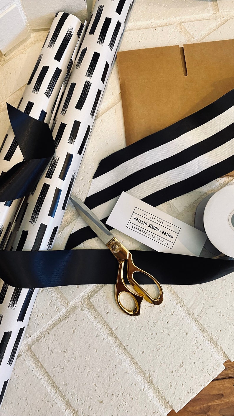 Op de afbeelding: Zwart-wit gestreept lint, zwart lint, gouden schaar en een rol zwart-wit gepatroneerd inpakpapier. Een klein wit kaartje met de tekst "EST 2020 KATELIN SIMONS design HANDMADE WITH LOVE XO" ligt op het lint.