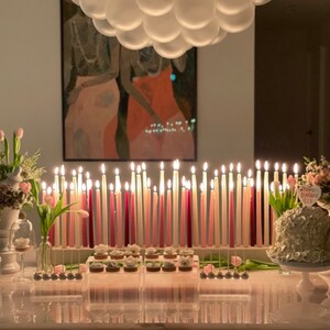 Op de afbeelding: Een tafeldecoratie voor een verjaardagsfeest met een witte taart versierd met een roze "Happy Birthday" bord, roze en witte kaarsen en roze en witte bloemen.