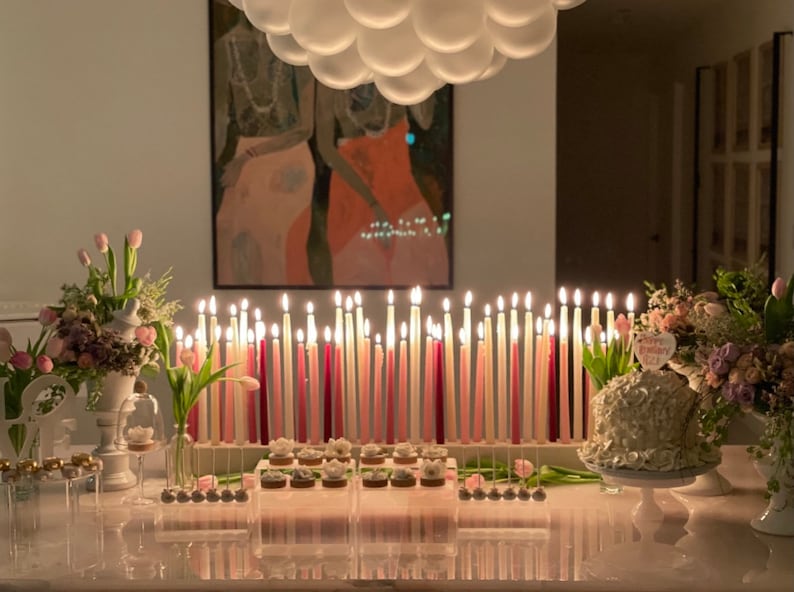 Op de afbeelding: Een tafeldecoratie voor een verjaardagsfeest met een witte taart versierd met een roze "Happy Birthday" bord, roze en witte kaarsen en roze en witte bloemen.