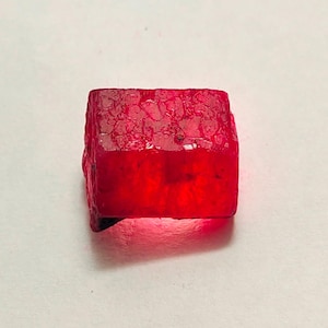 Puede incluir: Una gema de rubí en bruto, de forma rectangular y color rojo intenso. La parte superior tiene un aspecto texturizado y cristalino, mientras que la parte inferior es más translúcida. La gema está sobre un fondo blanco.