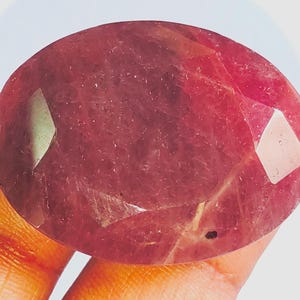 May include: A faceted, oval-cut ruby gemstone. The ruby displays a rich, deep red hue with internal inclusions. The facets catch the light, enhancing the gem's sparkle. The gemstone is held in a hand.