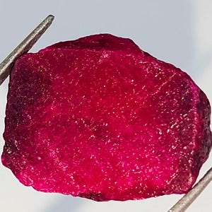 May include: A deep red ruby gemstone with a rough, natural finish. The ruby is a vibrant, rich red color and has a slightly irregular shape.