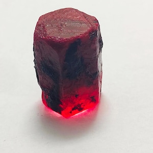 Puede incluir: Un rubí en bruto sin cortar. La gema es de color rojo intenso con inclusiones oscuras, casi negras. La parte inferior de la piedra brilla con una luz roja brillante. La piedra tiene una superficie rugosa y texturizada y forma rectangular.