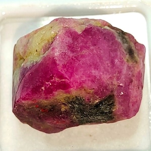 May include: A rough, pink ruby gemstone with black and green inclusions. The gemstone is in a white square display box.