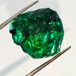 May include: A large, rough, green emerald gemstone. The emerald is held by a pair of tweezers.
