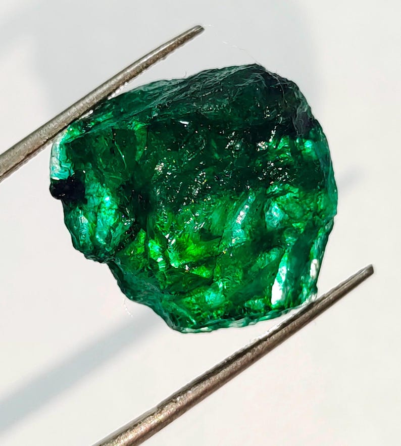 May include: A large, rough, green emerald gemstone. The emerald is held by a pair of tweezers.